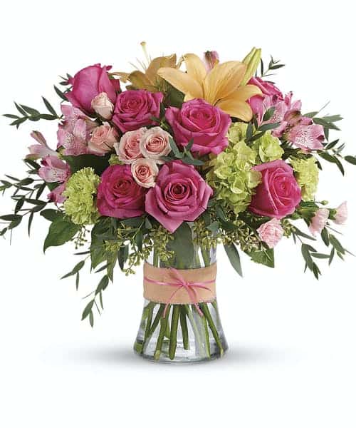 Put a spring in their step with this beautifully blushing bouquet of hot pink roses, soft peach lilies and fresh green hydrangea. Arranged in a graceful vase tied with a charming bow, it's a chic treat for any occasion!